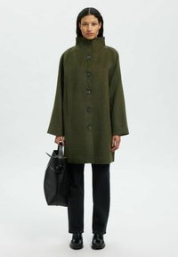 Groene wollen jas met een hoge kraag, grote zwarte knopen en zijzakken, gecombineerd met donkere jeans en een zwarte tas.