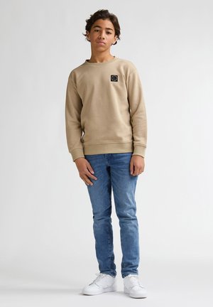 Beige sweatshirt met ronde hals, zwarte logopatch op de borst; gecombineerd met een blauwe spijkerbroek en witte sneakers. Glad textuur, casual ontwerp.