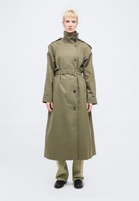 Άτομο που φοράει μακρύ χακί παλτό με ζώνη πάνω από ταιριαστό παντελόνι και μαύρα παπούτσια, στέκεται μπροστά σε μια απλή φωτεινή επιφάνεια.