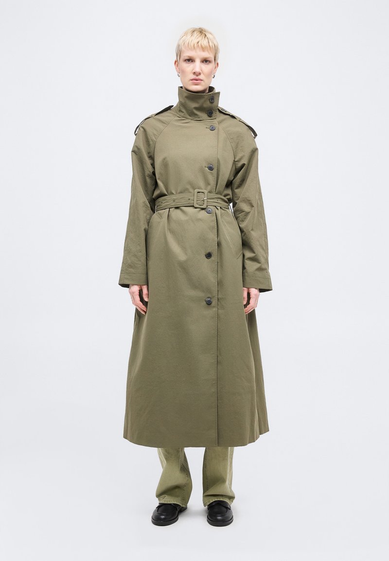 Άτομο που φοράει μακρύ χακί παλτό με ζώνη πάνω από ταιριαστό παντελόνι και μαύρα παπούτσια, στέκεται μπροστά σε μια απλή φωτεινή επιφάνεια.