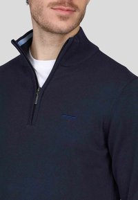 Pull-over marine avec col zippé, fabriqué en tissu tricot doux. Présente un col intérieur bleu clair et un logo brodé subtil sur la poitrine.