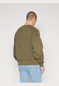 Felpa verde oliva dalla vestibilità rilassata, con polsini e orlo a coste, caratterizzata da un classico scollo tondo e un dettaglio triangolare sul retro dello scollo.