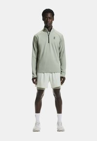 Hellgrüner Sweatshirt mit Viertel-Reißverschluss und Logo, kombiniert mit weißen Shorts. Glatte Textur, sportliche Passform und minimale Designelemente.