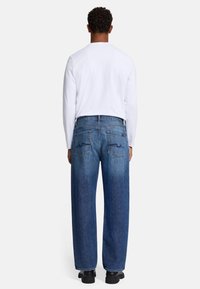 Jeans in denim blu con un design a gamba dritta, due tasche posteriori e delicate cuciture decorative, abbinati a una camicia bianca a maniche lunghe.