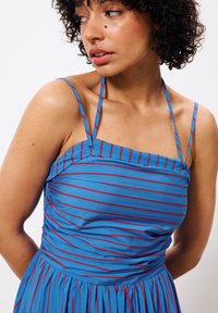 Trägerloses blaues Kleid mit roten horizontalen Streifen, strukturierter Stoff. Verfügt über ein gerafftes Oberteil und dünne Träger sowie einen fließenden, plissierten Rock.