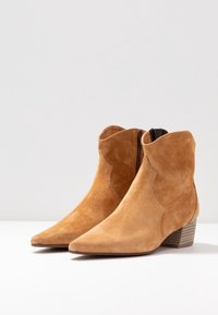 Botas de tobillo de ante color tostado con punta afilada, bajo tacón apilado y cremallera lateral. Presentan un borde superior con ondas y una textura suave.