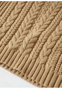 Tissu beige tricoté avec des motifs de câbles tressés épais et un ourlet côtelé, montrant une texture détaillée et un travail de points.