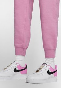 Vita Nike Air Force 1-sneakers med rosa accenter och svart swoosh. Skon har metalliska öglor och texturerade snören. Kombinerat med rosa sweatpants.