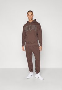Moletom de algodão castanho com gráfico "NIKECOURT", combinado com calças de treino à medida. Ténis brancos completam o visual atlético.