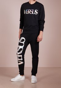 Sudadera negra con "VERSUS" en letras iridiscentes, combinada con pantalones joggers negros que presentan un gran texto iridiscente "SNEAKERS" y zapatos blancos.