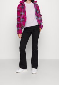 Veste en polaire rose à carreaux avec capuche, portée sur un pull violet clair et un pantalon noir évasé. Des baskets blanches complètent la tenue.