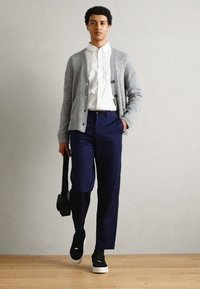 Grå strikket cardigan, hvit skjorte med krage, marineblå bukser, svarte joggesko. Holder en svart bag. Enkelt design, nøytrale farger, uformelt utseende.