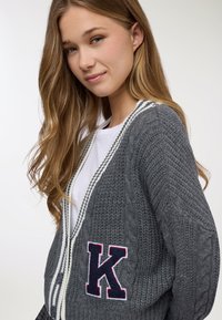 Giovane donna con capelli biondi lunghi che indossa un cardigan lavorato a maglia grigio con strisce bianche e una grande toppa a forma di "K" sopra una camicia bianca.