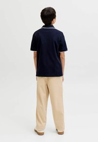 Garçon debout de dos, portant un polo bleu marine, un pantalon beige et des chaussures blanches avec des semelles marron, sur un fond clair uni.