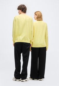 Άνδρας και γυναίκα στέκονται πλάι πλάι με την πλάτη προς τα εμπρός, και οι δύο φορούν κίτρινα πουλόβερ, μαύρο παντελόνι και λευκά αθλητικά παπούτσια, μπροστά σε ένα απλό λευκό φόντο.