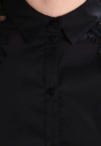 Blouse noire boutonnée avec col. Présente des détails texturés sur les épaules, fabriquée en tissu lisse avec une coupe ajustée.