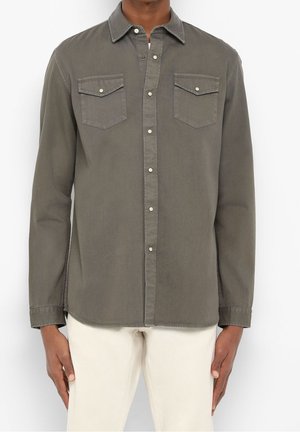 Mężczyzna w oliwkowo-zielonej koszuli na guziki z długim rękawem i kieszeniami z klapkami na piersi oraz beżowych spodniach, stojący na białym tle.