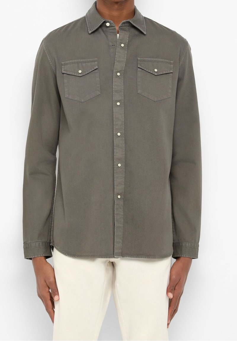 Homme portant une chemise à manches longues olive verte avec des poches poitrine à rabat et un pantalon beige, debout devant un fond blanc.