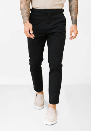 Pantaloni negri, tip slim, cu textură fină, prevăzuți cu bucle pentru curea și talie medie. Asortați cu încălțăminte slip-on de culoare deschisă.