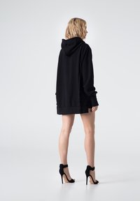 Czarna, oversize bluza z kapturem i trokiem, z boku rozcięcia, w połączeniu z czarnymi szpilkami. Modelka pokazana z tyłu.