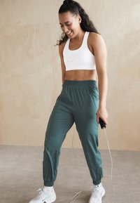 Witte sportbeha met een grijs logo, groene joggers met elastische boorden en zijzakken, en witte sportschoenen, terwijl ze een springtouw vasthoudt.