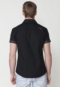 Camisa negra de manga corta con cuello, fabricada en un tejido ligero, con puños de mezclilla en contraste y un sutil logotipo en la parte de atrás.