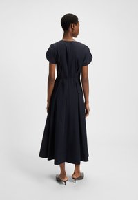 Žena s krátkymi vlasmi, oblečená v čiernych midi šatách s nafúknutými rukávmi a mules s nízkym podpätkom, stojaca chrbtom k jednoduchému pozadiu.