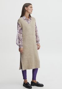 Abito beige lavorato a maglia senza maniche con scollatura a V, fessure laterali, indossato sopra una camicetta floreale viola e leggings viola, abbinato a mocassini neri.