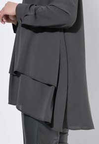 Person wearing layered dark gray loose-fitting top with buttoned cuff and black leather pants standing against a light background.