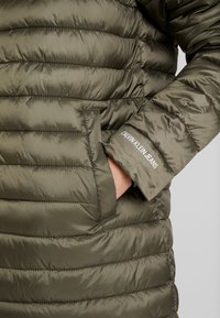 Olivově zelená zimní bunda s horizontálním prošíváním, boční kapsou a bílým nápisem "CALVIN KLEIN JEANS" na manžetě.