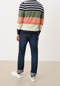 Mann trägt dunkelblaue, gekrempelte Jeans, weiße Sneakers und ein langärmliges, gestreiftes Shirt in mehreren Farben, steht vor einem einfarbigen Hintergrund.