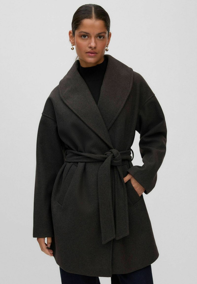 Femme portant un manteau en laine foncé ceinturé avec un col large, une main dans la poche et des boucles d'oreilles créoles dorées sur un fond clair uni.