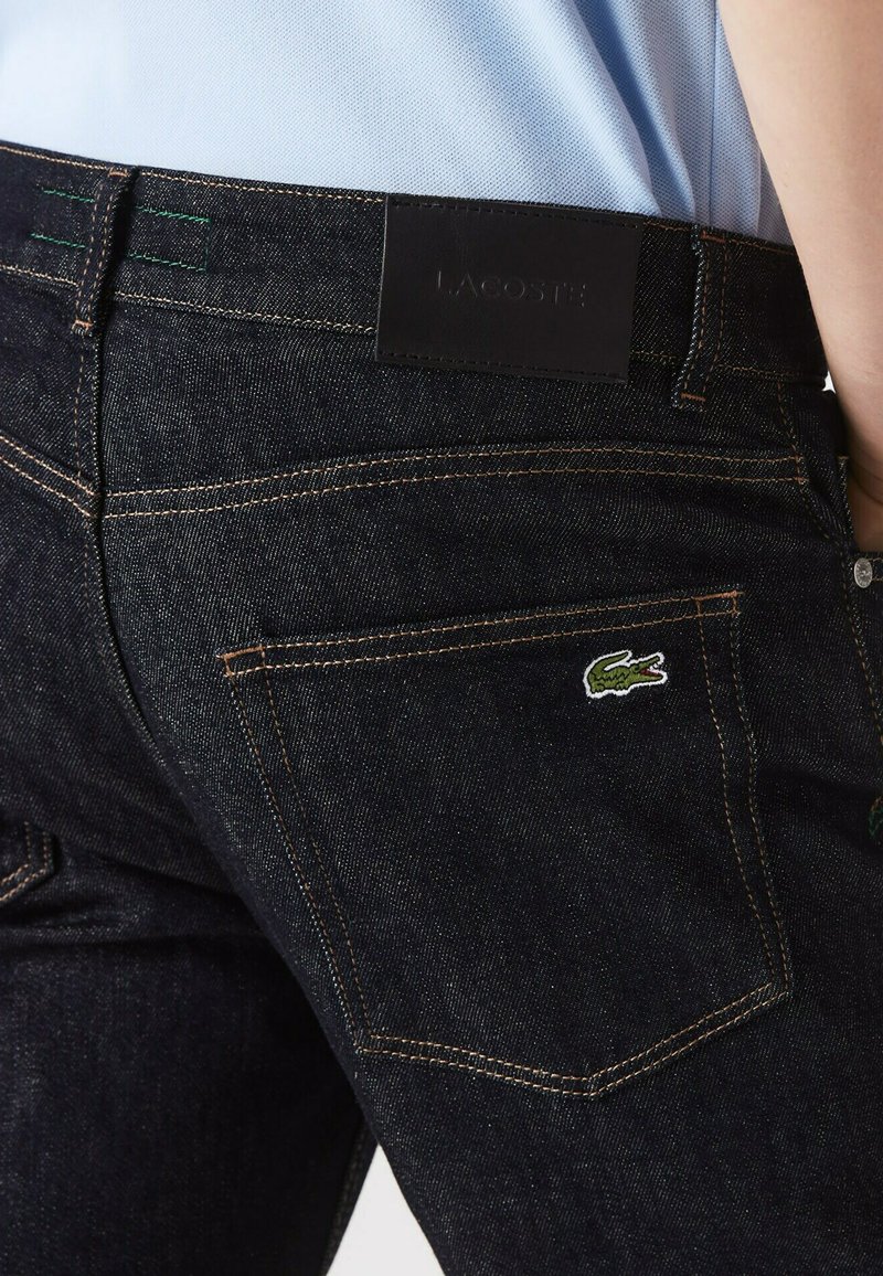 Person trägt dunkelblaue Jeans mit einem grünen, gestickten Krokodil-Logo auf der Gesäßtasche und einem schwarzen Lacoste-Label am Bund.