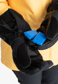 Des gants isolants noirs avec une texture lisse et des paumes rembourrées tiennent deux batteries bleues. Une veste jaune est partiellement visible en arrière-plan.