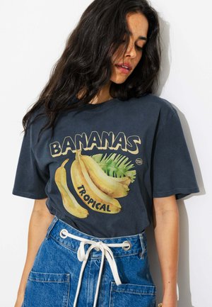 Femme aux longs cheveux foncés portant un T-shirt bleu foncé avec un motif banane jaune et les mots "Bananas Tropical", portée avec un jean bleu.