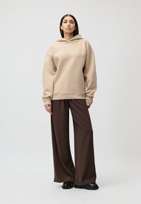Beige oversized hettegenser med snøring i hetten, kombinert med brune vide bukser. Dette antrekket er laget av glatt, noe teksturert stoff.