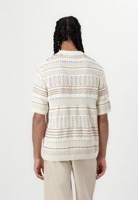 Pull en tricot crème avec des rayures horizontales brunes, à manches courtes et à maille ouverte. Texture douce avec des motifs et des accents variés.