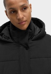 Giacca a vento nera con colletto alto e cappuccio regolabile. Caratterizzata da chiusure a bottoni e texture trapuntata. Materiale leggero.