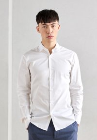 Weißes Langarm-Hemd mit Knopfverschluss und klassischem Kragen, glatter Textur und Slim-Fit. Das Model steht vor einem neutralen Hintergrund.