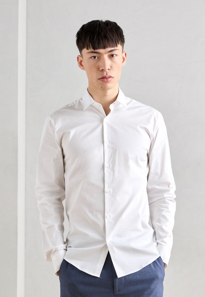 Witte langemouwen knopshirt met een klassieke kraag, een gladde textuur en een slim fit. Model staat tegen een neutrale achtergrond.
