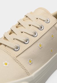 Μπεζ καμβαδένιο αθλητικό παπούτσι με κρέμ κορδόνια και μικρές λευκές κεντημένες μαργαρίτες με κίτρινο κέντρο στο πλάι.