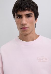 Nuori mies, jolla on tummat kiharat hiukset, pukeutuneena vaaleanpunaiseen collegepaitaan, jossa on "Scalpers Studio House of Antiques" -logo vaaleanharmaata taustaa vasten.