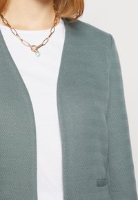 Texturovaný světle zelený blazer s vzorem herringbone, s klopovým límcem a otevřeným předním dílem, navlečený přes bílý tričko.