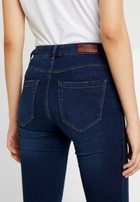 Jeans de mezclilla azul oscuro con un diseño de cintura alta, que cuentan con bolsillos traseros y una etiqueta de cuero en la cinturilla. Textura suave, corte ajustado.