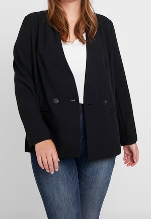 Kvinde iført en sort knappet blazer over en hvid skjorte og blå jeans, stående mod en ensfarvet lysegrå baggrund.