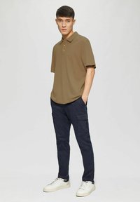 Khakifarbene Polo-Shirts mit einer Knopfleiste mit drei Knöpfen, kombiniert mit dunkelblauen Cargo-Hosen. Das Modell trägt weiße Turnschuhe und steht vor einem neutralen Hintergrund.