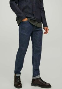 Veste en denim et jeans bleu foncé avec une chemise verte en dessous. Les jeans ont les poignets retroussés et la tenue est complétée par des bottes marron à lacets.