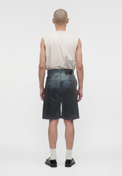 Ένα άτομο με ξυρισμένο κεφάλι φοράει ένα μπεζ αμάνικο πουκάμισο, ξεθωριασμένα τζιν σορτς μέχρι το γόνατο, λευκές κάλτσες και μαύρα παπούτσια, στέκεται γυρισμένο προς τα πίσω.