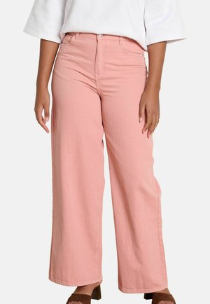 Osoba oblečená v ružových nohaviciach s vysokým pásom a širokými nohavicami, v kombinácii s bielym tričkom a hnedými sandálmi na podpätku s otvorenou špičkou.