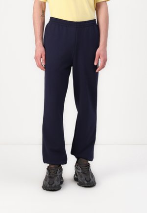 Homme portant un pantalon de sport noir, un t-shirt jaune et des baskets noires, debout devant un fond blanc uni.
